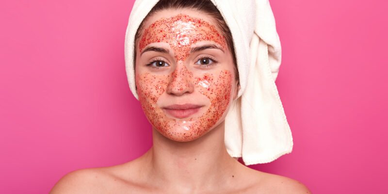 young-attractive-woman-with-white-towel-on-her-head-has-naked-body-smilling-isolated-over-pink-wall-in-studio-looks-directly-at-camera-having-red-scrub-on-her-face